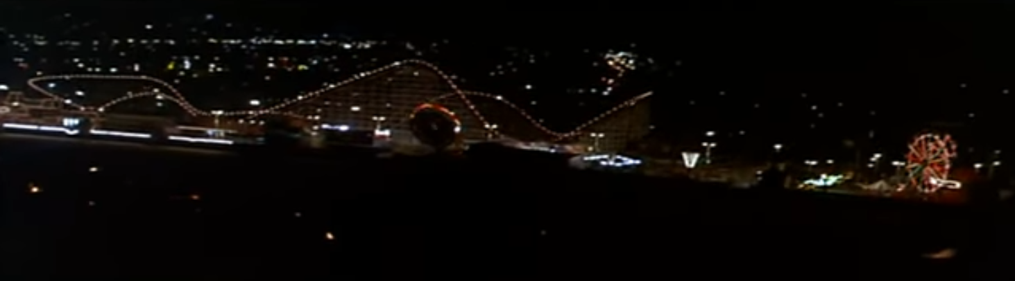 Aerial view of Giant Dipper from the opening shot of The Lost Boys, its night time, the track is lit with string lights. There are also other flat rides lit up along the rest of the boardwalk