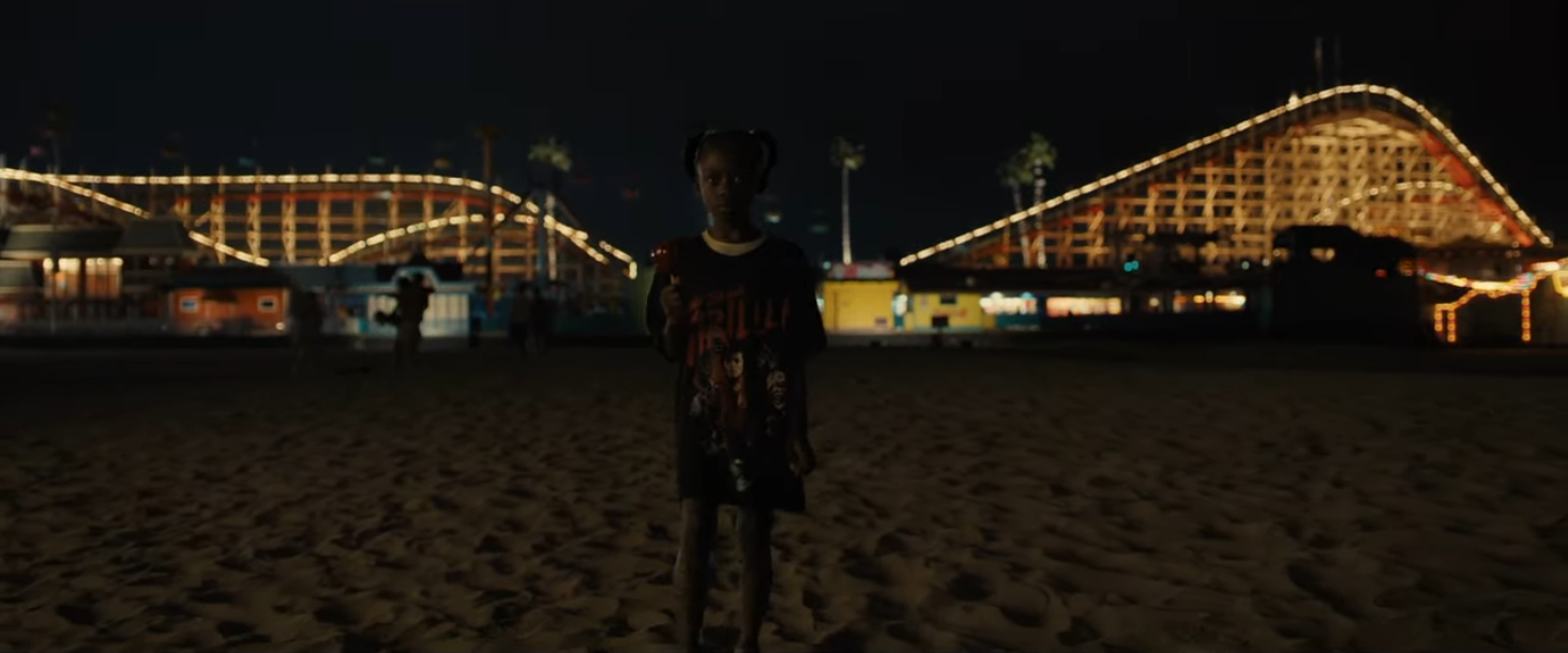 Adilade standing on a beach holding a red candied apple, in the background, out of focus, Giant Dipper towers over, its track is lit by string lights.
