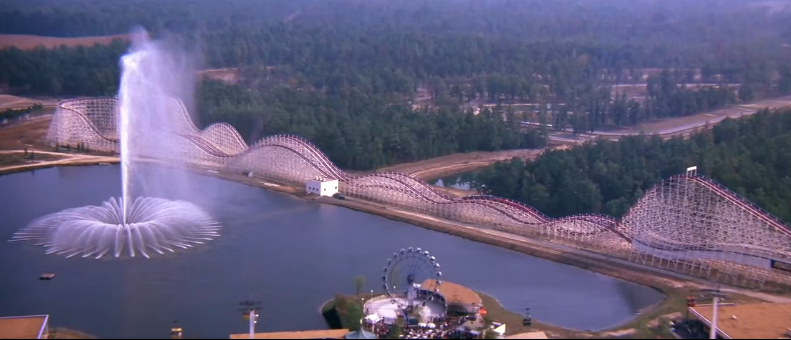 Aerial view of Racer 75 in the movie Rollercoaster from the side, it's painted white and its next to a very large lake.